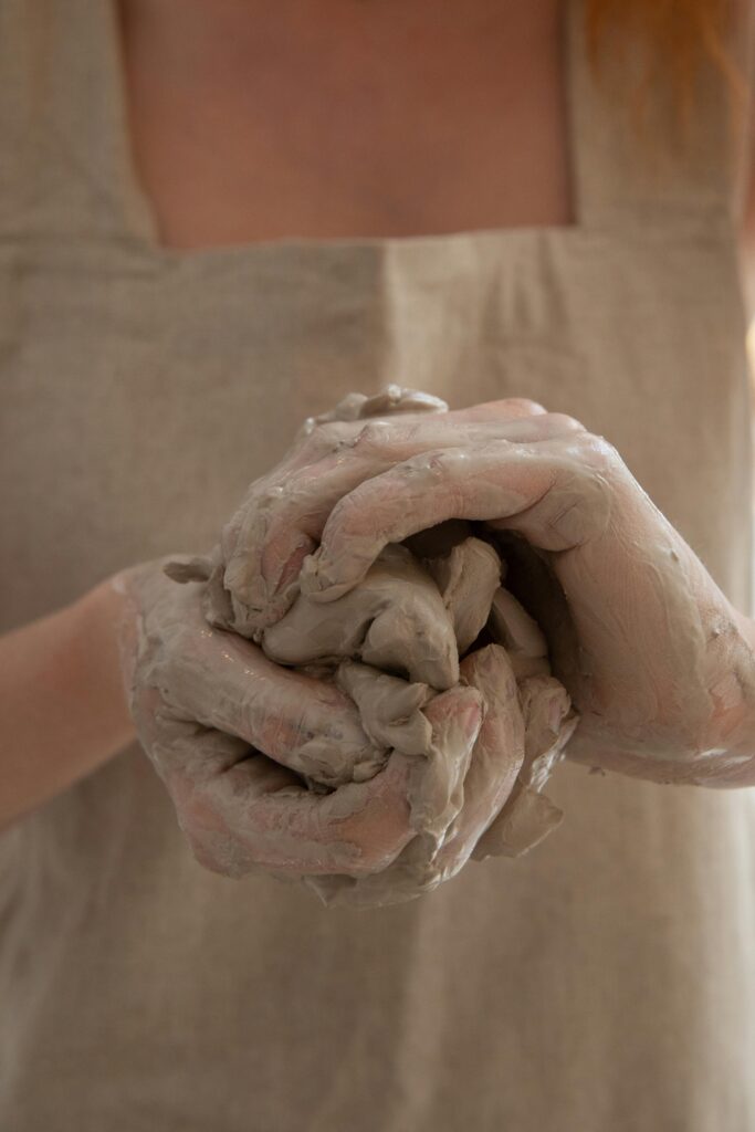 Unrecognizable person wearing gray apron shaping and molding clay in dirty hands while making handicraft crockery in workshop during work